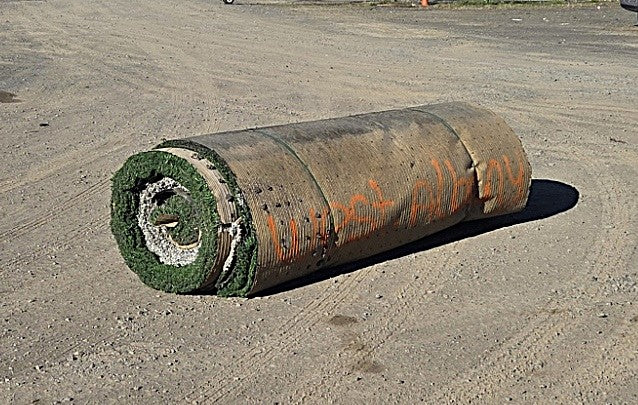 Rolled-up used artificial turf resting on dirt, showing the ribbed backing with "West Albany" written in orange spray paint