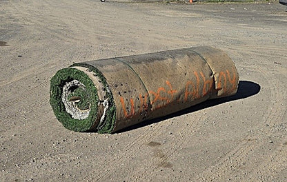 Rolled-up used artificial turf resting on dirt, showing the ribbed backing with "West Albany" written in orange spray paint