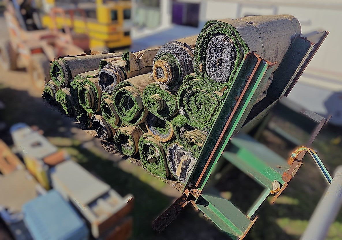 Multiple rolls of salvaged artificial turf stacked on a large green metal forklift attachment rack