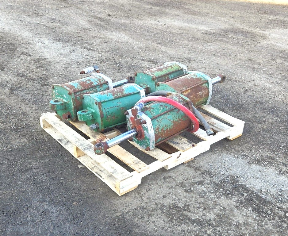 Four green heavy-duty pneumatic tie-rod cylinders resting on a wooden pallet showing swivel eye rod ends and connecting hoses.
